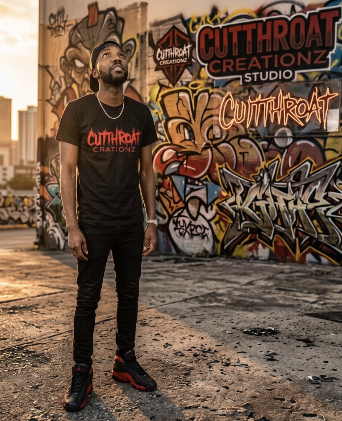 Cutthroat Creationz founder in a black flame-logo tee in front of the Cutthroat Creationz graffiti studio wall